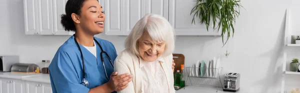 Enfermera multirracial feliz en uniforme teniendo cuidado de la mujer mayor alegre en casa, pancarta - foto de stock