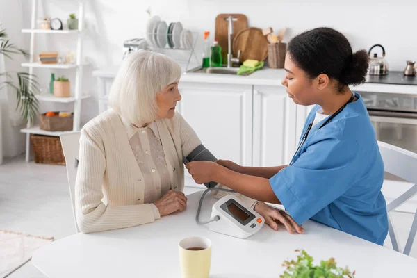 Enfermera multirracial que mide la presión arterial de la mujer mayor con pelo gris - foto de stock
