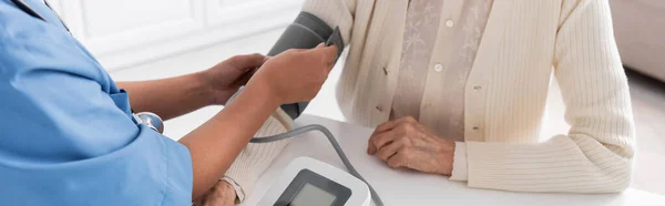 Cropped view of multiracial nurse measuring blood pressure of senior woman, banner — Stock Photo