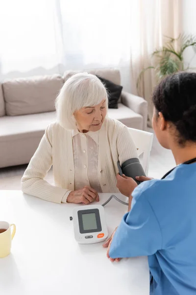 Enfermera multirracial que mide la presión arterial de una mujer jubilada con pelo gris - foto de stock
