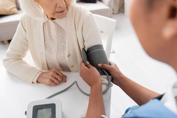 Partial view of multiracial nurse measuring blood pressure of senior woman with grey hair — Stock Photo