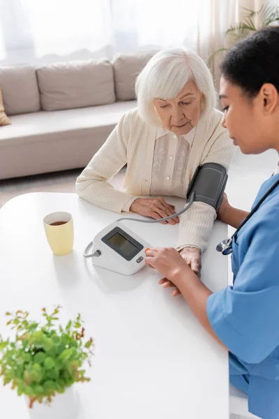 Cuidador multirracial medir a pressão arterial de mulher idosa com cabelos grisalhos — Fotografia de Stock