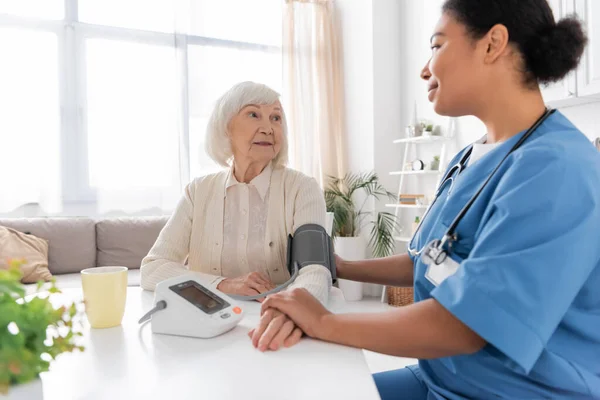 Весела багаторасова медсестра вимірює кров'яний тиск старшої жінки з сірим волоссям — стокове фото
