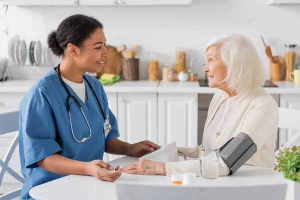 Happy multiracial nurse measuring blood pressure of senior woman with grey hair next to medication on table — Stock Photo