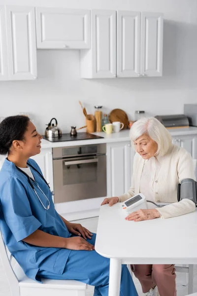 Infirmière multiraciale heureuse regardant la femme âgée avec les cheveux gris mesurant la pression artérielle — Photo de stock