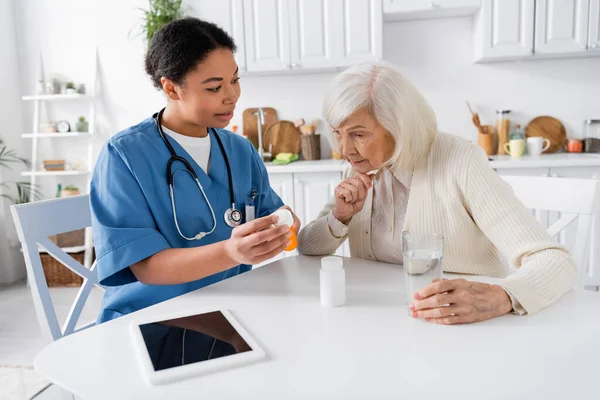 Многорасовая медсестра показывает лекарства пожилой женщине рядом с цифровым планшетом на столе — стоковое фото