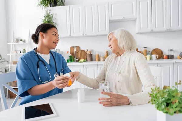 Багаторасовий медсестра показує ліки скептично старшій жінці поруч з цифровим планшетом на столі — стокове фото