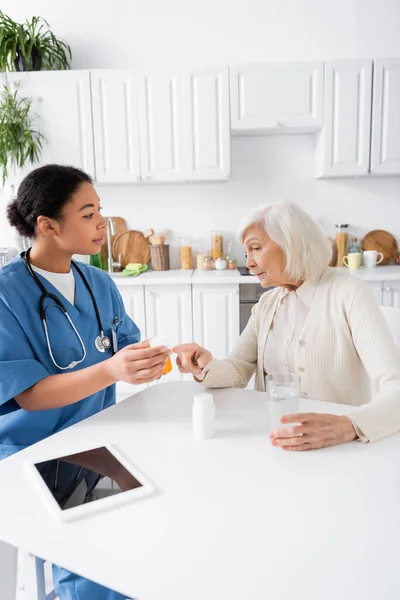 Старша жінка вказує пальцем на таблетки в руках багаторасової медсестри — стокове фото