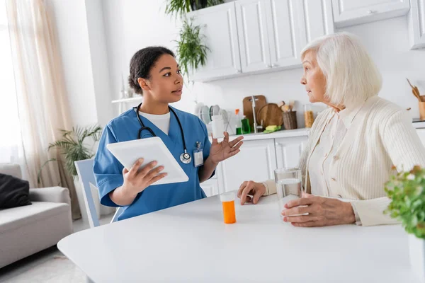 Infirmière multiraciale brune utilisant une tablette numérique et tenant des médicaments tout en parlant à une femme âgée aux cheveux gris à la maison — Photo de stock