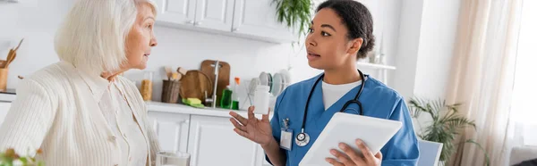 Brunette multiracial nurse using digital tablet and holding medication while talking to senior woman with grey hair, banner — Stock Photo