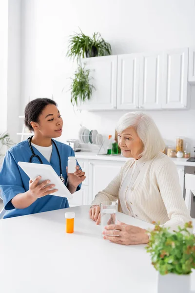 Brünette multirassische Krankenschwester mit digitalem Tablet und Medikamenten in der Hand, während sie mit einer Rentnerin mit grauen Haaren spricht — Stockfoto