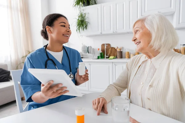 Glückliche multiethnische Krankenschwester mit digitalem Tablet und Medikamenten in der Hand, während sie mit einer Rentnerin mit grauen Haaren spricht — Stockfoto