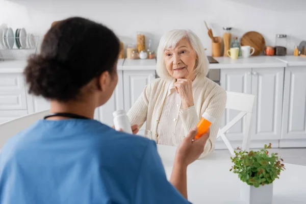 Женщина в отставке с седыми волосами смотрит на многорасовую медсестру с лекарствами на размытом переднем плане — стоковое фото