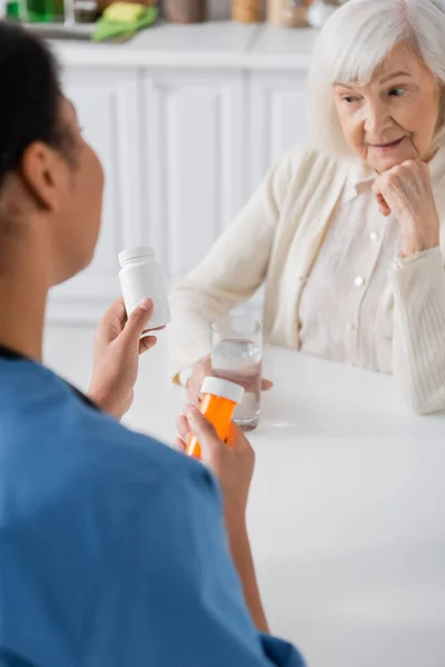 Пенсіонерка з сірим волоссям дивиться на ліки в руках багаторасової медсестри на розмитому передньому плані — стокове фото