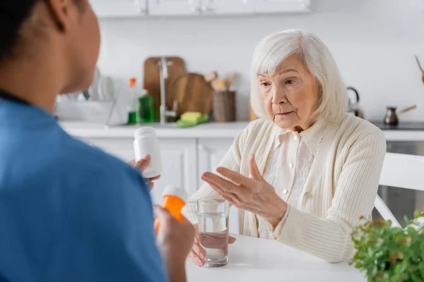 Пенсіонерка з сірим волоссям запитує про ліки в руках багаторасової медсестри на розмитому фоні — стокове фото