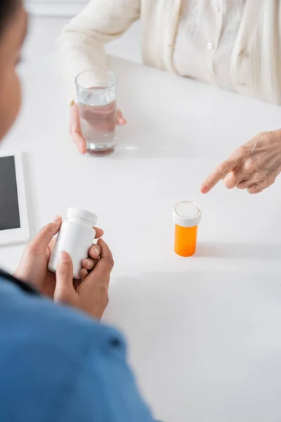 Cropped view of retired woman pointing at bottle with medication near multiracial nurse on blurred foreground — Stock Photo