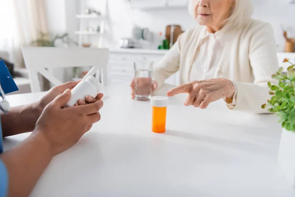 Visão cortada de mulher idosa apontando para garrafa com medicação perto de enfermeira multirracial em primeiro plano turvo — Fotografia de Stock