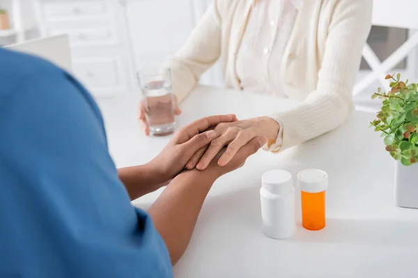 Teilansicht einer multiethnischen Krankenschwester, die Hand hält, während sie eine Seniorin neben Medikamenten auf dem Tisch tröstet — Stockfoto