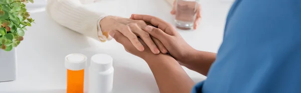 Visão cortada de enfermeira multirracial segurando a mão enquanto conforta a mulher idosa ao lado de medicação na mesa, banner — Fotografia de Stock