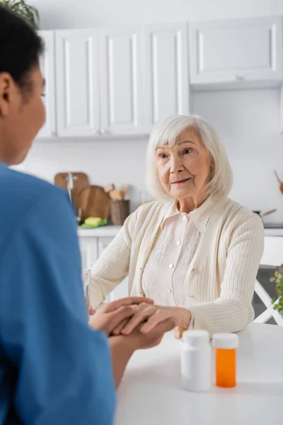 Enfermera multirracial borrosa cogida de la mano mientras reconforta a la mujer mayor junto a la medicación en la mesa - foto de stock