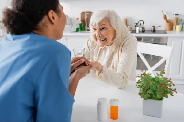 Infirmière multiraciale tenant la main avec une femme âgée heureuse à côté de médicaments sur la table — Photo de stock