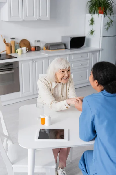 Multiracial nurse holding hands with cheerful senior woman next to medication on table — Stock Photo