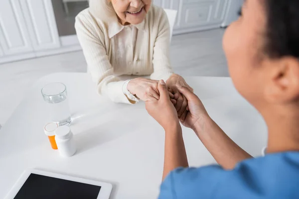 Blurred multiracial nurse holding hands with positive senior woman next to medication on table — Stock Photo