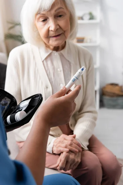Multiracial caregiver holding lancet pen near senior woman with grey hair — Stock Photo