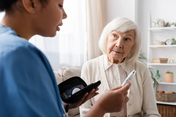 Enfermera multirracial que sostiene la pluma de la lanceta y explica cómo utilizarla a la mujer mayor con pelo gris - foto de stock