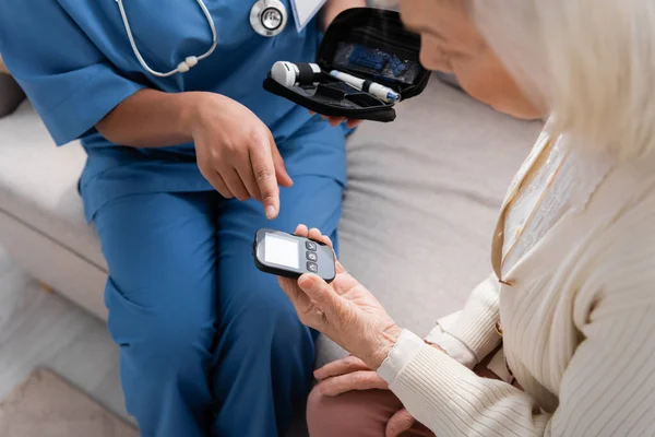 Multiracial nurse holding diabetes kit and pointing at glucometer near senior woman — Stock Photo