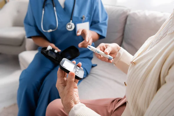 Abgeschnittene Ansicht einer Seniorin mit Lanzetstift und Glukometer in der Nähe einer Krankenschwester, die auf dem Sofa sitzt — Stockfoto