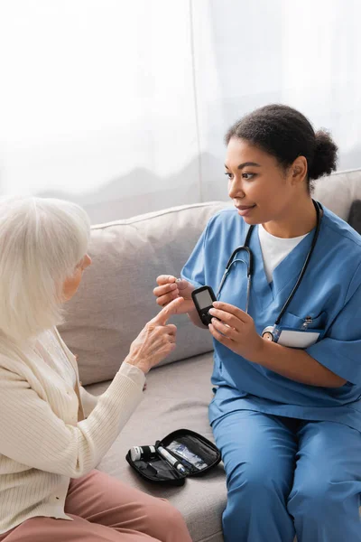 Брюнетка многорасовая медсестра в форме проведение тест-полоса и глюкометр рядом со старшей женщиной — стоковое фото
