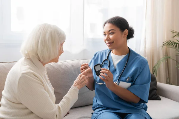 Happy multiracial nurse in uniform holding test strip and glucometer near senior woman — Stock Photo
