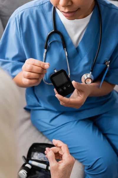 Ausgeschnittene Ansicht einer multirassischen Krankenschwester in Uniform, die Glukometer mit Teststreifen in der Nähe einer Seniorin hält — Stockfoto