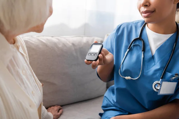 Ausgeschnittene Ansicht einer multirassischen Pflegerin in Uniform mit Glukometer in der Nähe einer Seniorin mit grauen Haaren — Stockfoto