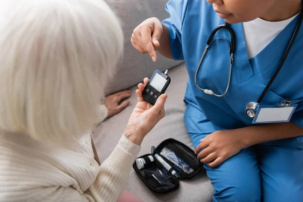 Ausgeschnittene Ansicht einer multirassischen Pflegerin in Uniform, die auf ein Glukometer in der Nähe einer Seniorin mit grauen Haaren zeigt — Stockfoto