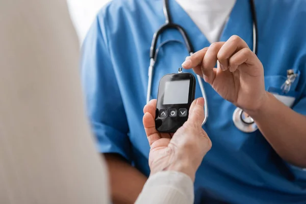Ausgeschnittene Ansicht einer multirassischen Krankenschwester, die mit dem Finger auf Glukometer mit Teststreifen in der Nähe einer Seniorin zeigt — Stockfoto