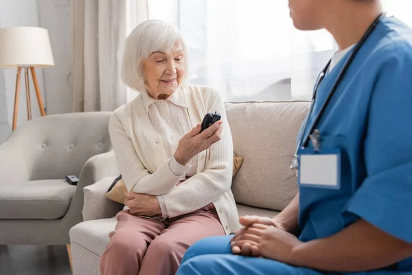 Счастливая пожилая женщина смотрит на глюкометр рядом с многорасовой медсестрой в синей форме — стоковое фото