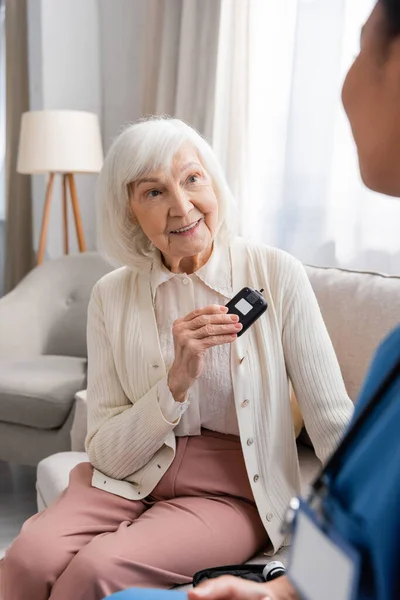Счастливая пожилая женщина с седыми волосами держит глюкометр рядом с многорасовой медсестрой в синей форме — стоковое фото