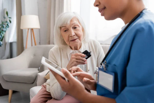 Mujer mayor con cabello gris sosteniendo glucosímetro cerca de enfermera multirracial con tableta digital - foto de stock