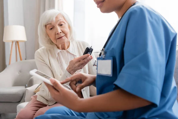 Пожилая женщина с седыми волосами держит глюкометр рядом с многорасовой медсестрой с помощью цифрового планшета — стоковое фото