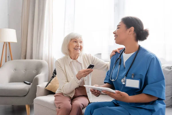 Joyeuse femme âgée avec les cheveux gris tenant glucomètre près infirmière multiraciale heureuse en utilisant une tablette numérique — Photo de stock