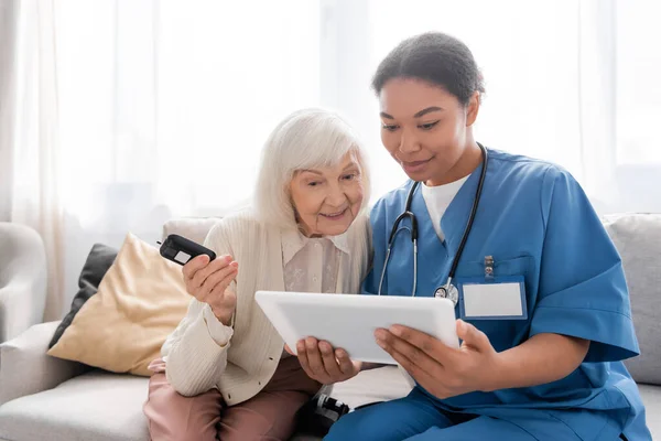 Веселая пожилая женщина с седыми волосами, держащая глюкометр и глядя на цифровой планшет около счастливой многорасовой медсестры — стоковое фото