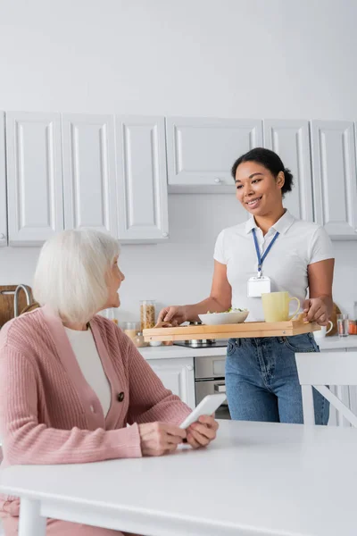 Щасливий багаторасовий соціальний працівник тримає піднос з обідом для старшої жінки з сірим волоссям — стокове фото