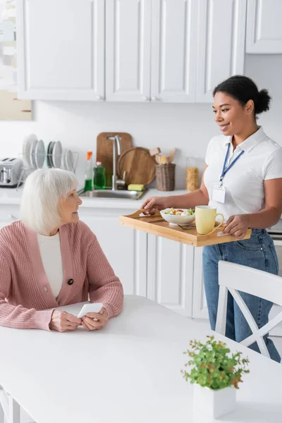 Alegre multirracial assistente social segurando bandeja com almoço para a mulher idosa com cabelos grisalhos — Fotografia de Stock