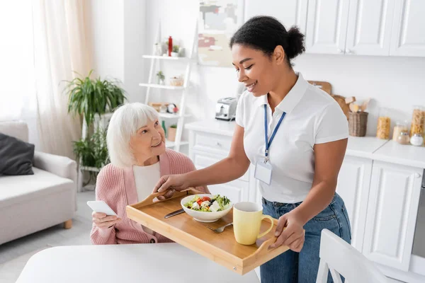 Happy multiracial social worker holding tray with lunch for senior woman with grey hair — Stock Photo