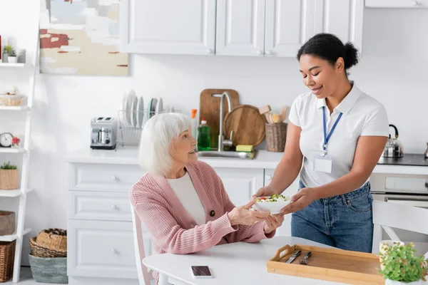 Felice assistente sociale multirazziale in possesso di ciotola con insalata per donna anziana con i capelli grigi — Foto stock