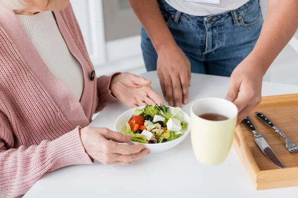 Cropped view of multiracial social worker serving lunch near senior woman — Stock Photo