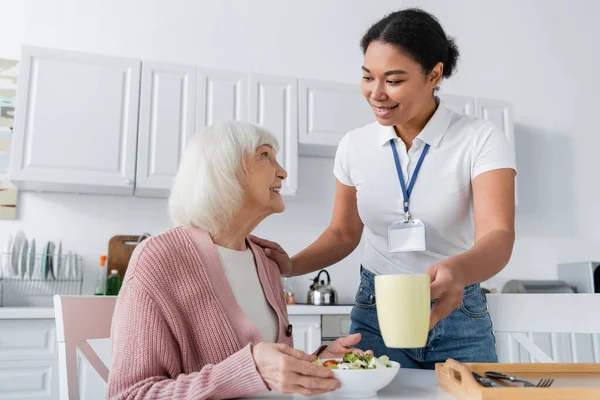Feliz trabajador social multirracial sirviendo el almuerzo a la sonriente mujer mayor con el pelo gris - foto de stock
