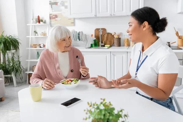 Happy senior woman with grey hair eating salad and talking with multiracial social worker — Stock Photo